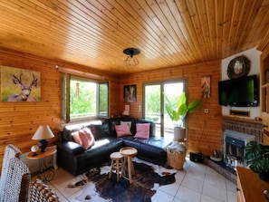 A log cabin interior with a black leather sofa, a television, a fireplace, and a large window.