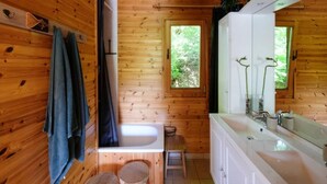 Baño de cabaña de madera con bañera, lavamanos y ventana con vista a follaje.