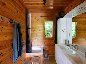 Baño de cabaña de madera con bañera, lavamanos y ventana con vista a follaje.