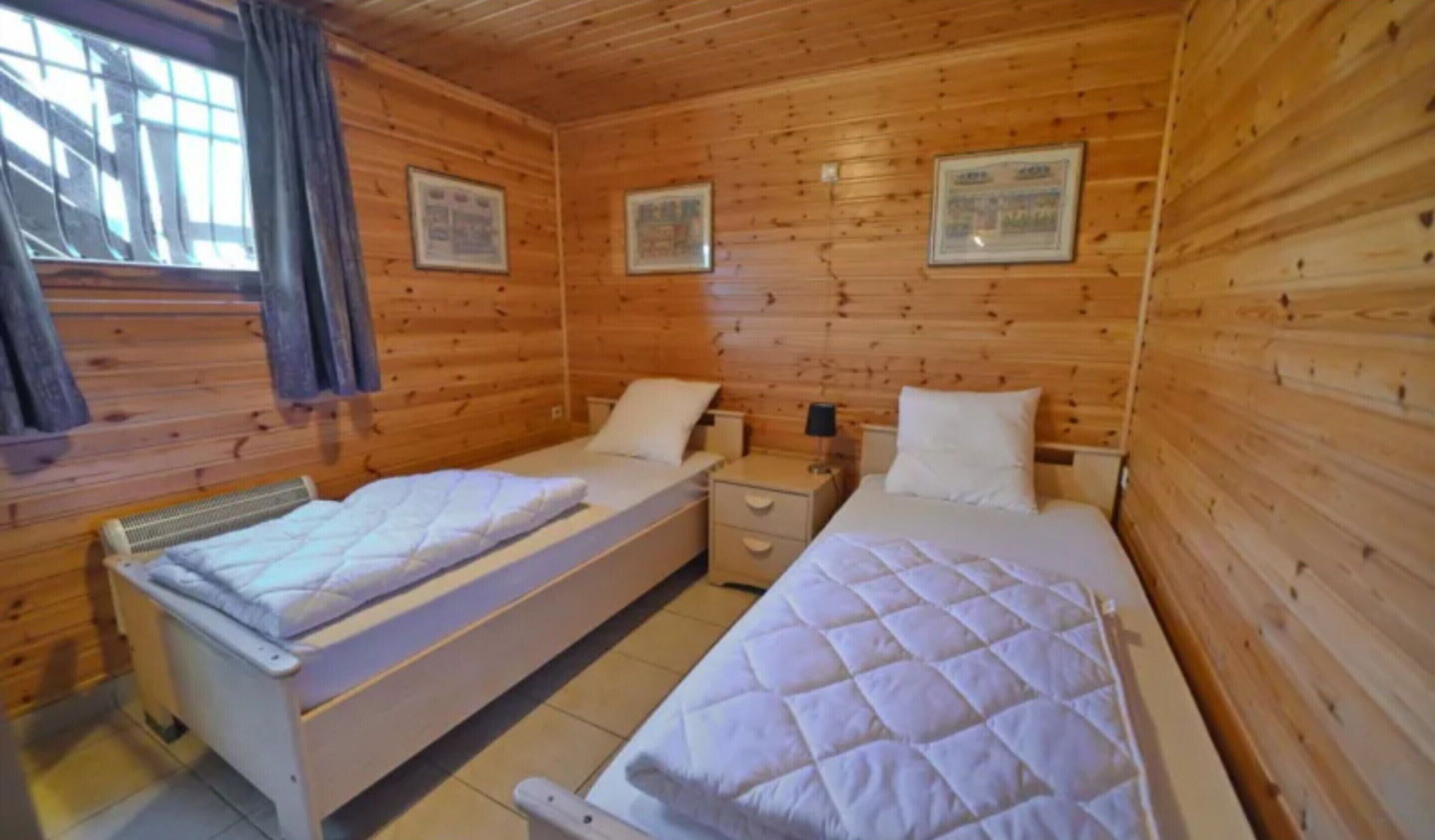 Ein Zimmer in einer Holzhütte mit zwei Betten, einem Fenster mit Vorhängen und gerahmten Bildern an den Wänden.