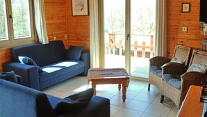 A cozy cabin interior with a blue sofa, wicker chairs, a wooden table, and a sliding glass door leading to an outdoor deck. - Holiday Home in La Roche With Sauna (La Roche-en-Ardenne)