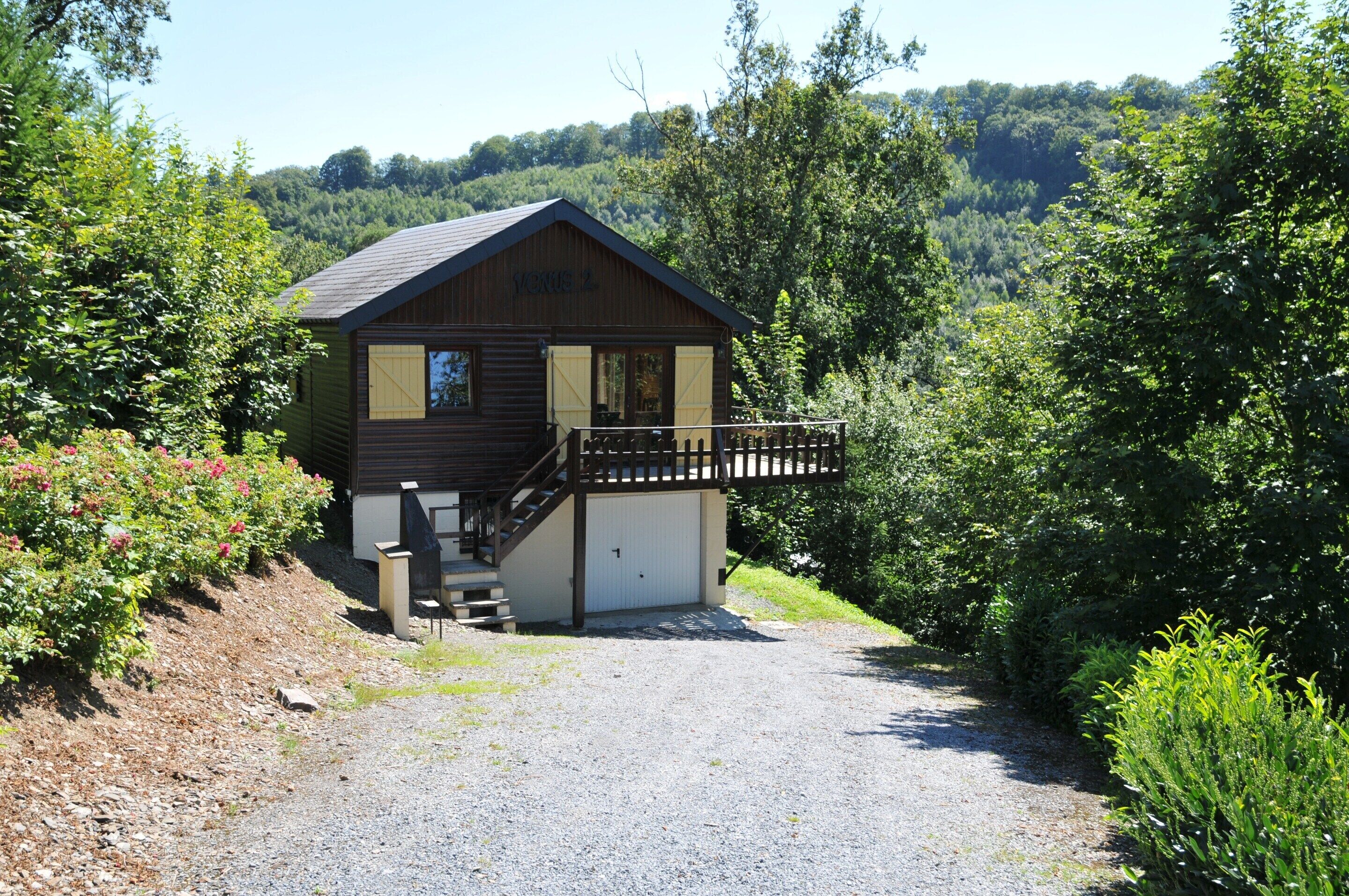 A wooden cabin with a porch, a garage, and a gravel driveway surrounded by greenery.