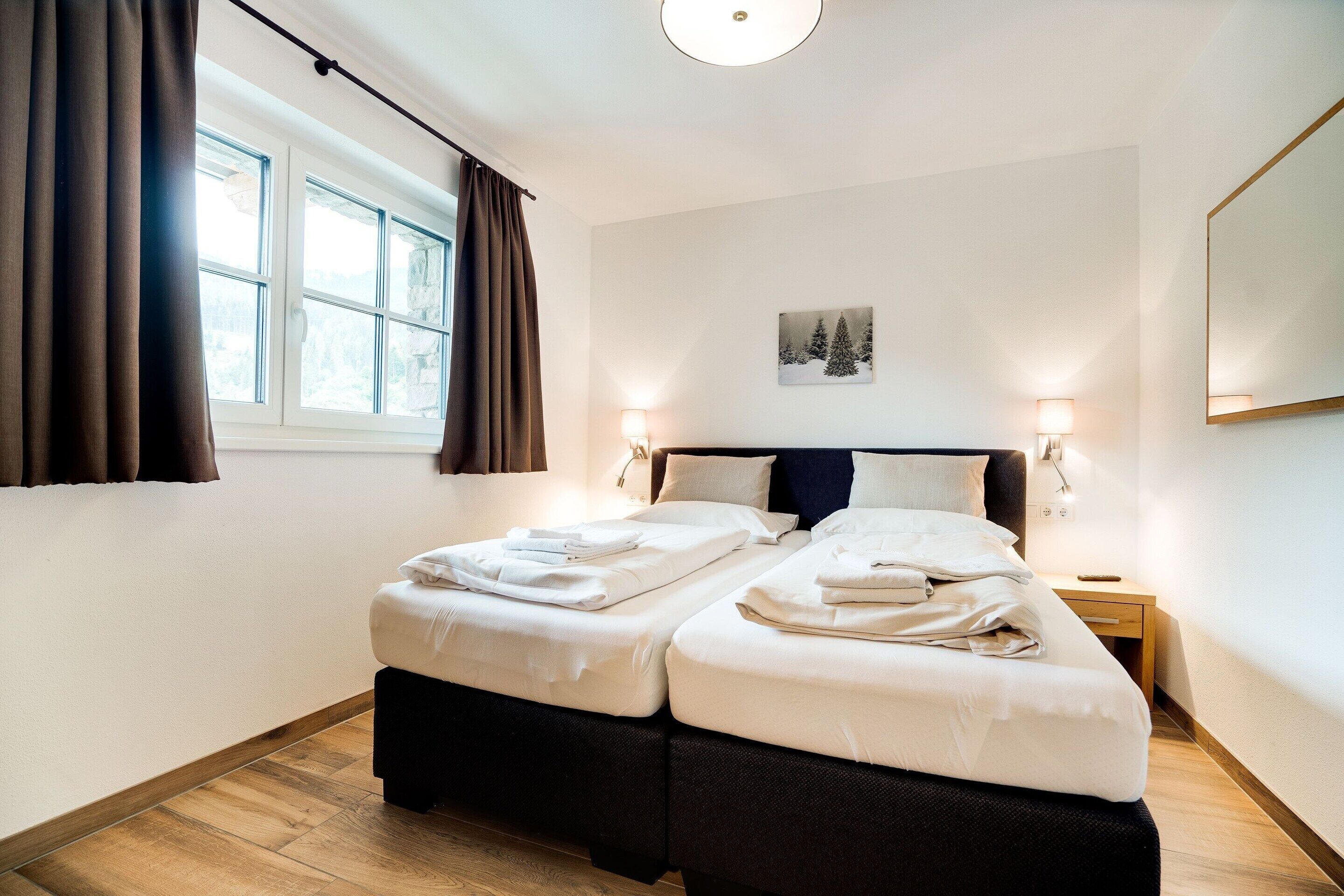 A hotel room with two beds, a window with curtains, a painting on the wall, and bedside lamps.