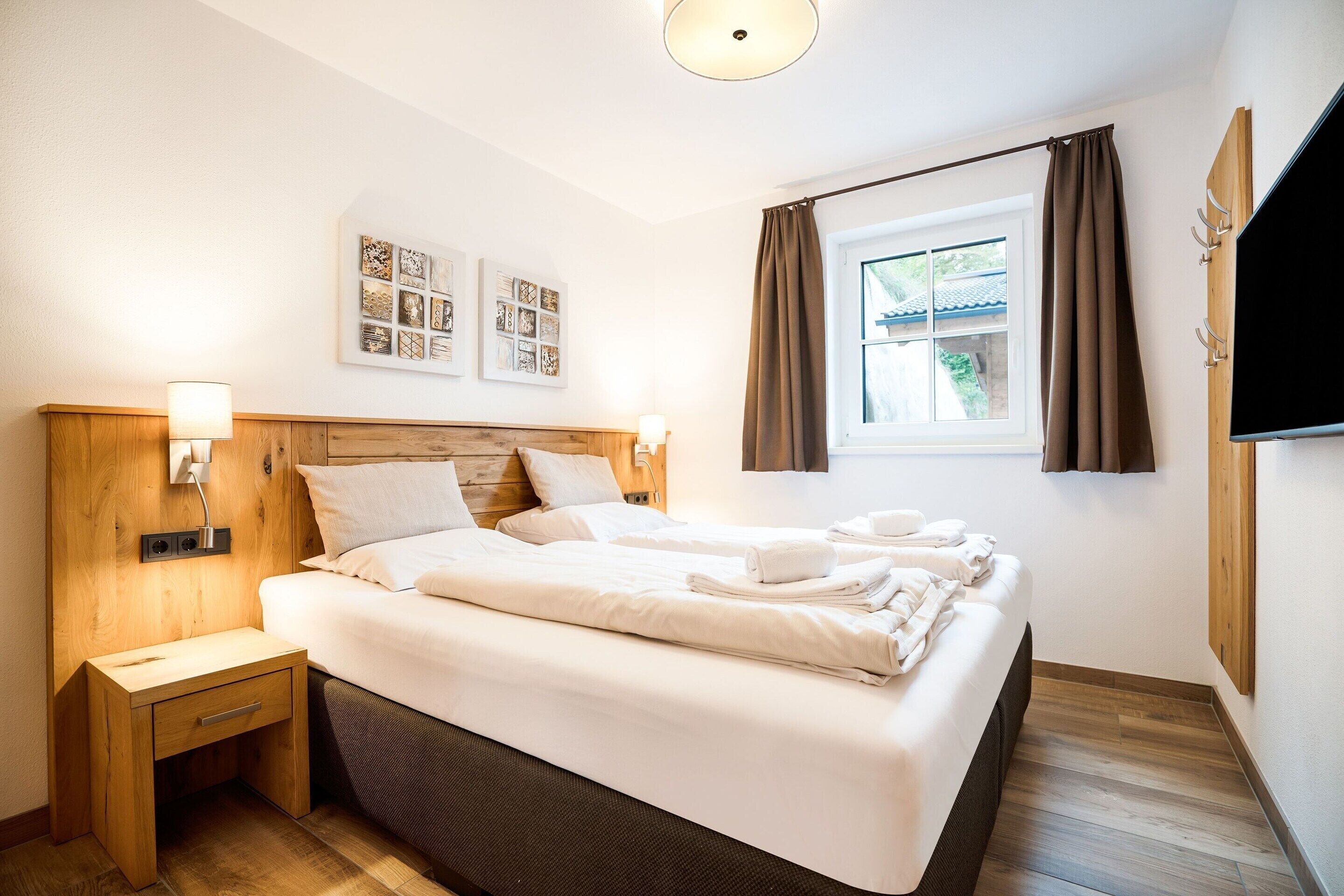 Une chambre d’hôtel avec un lit, des tables de chevet, des œuvres d’art au mur, une fenêtre avec des rideaux et un téléviseur à écran plat fixé au mur.