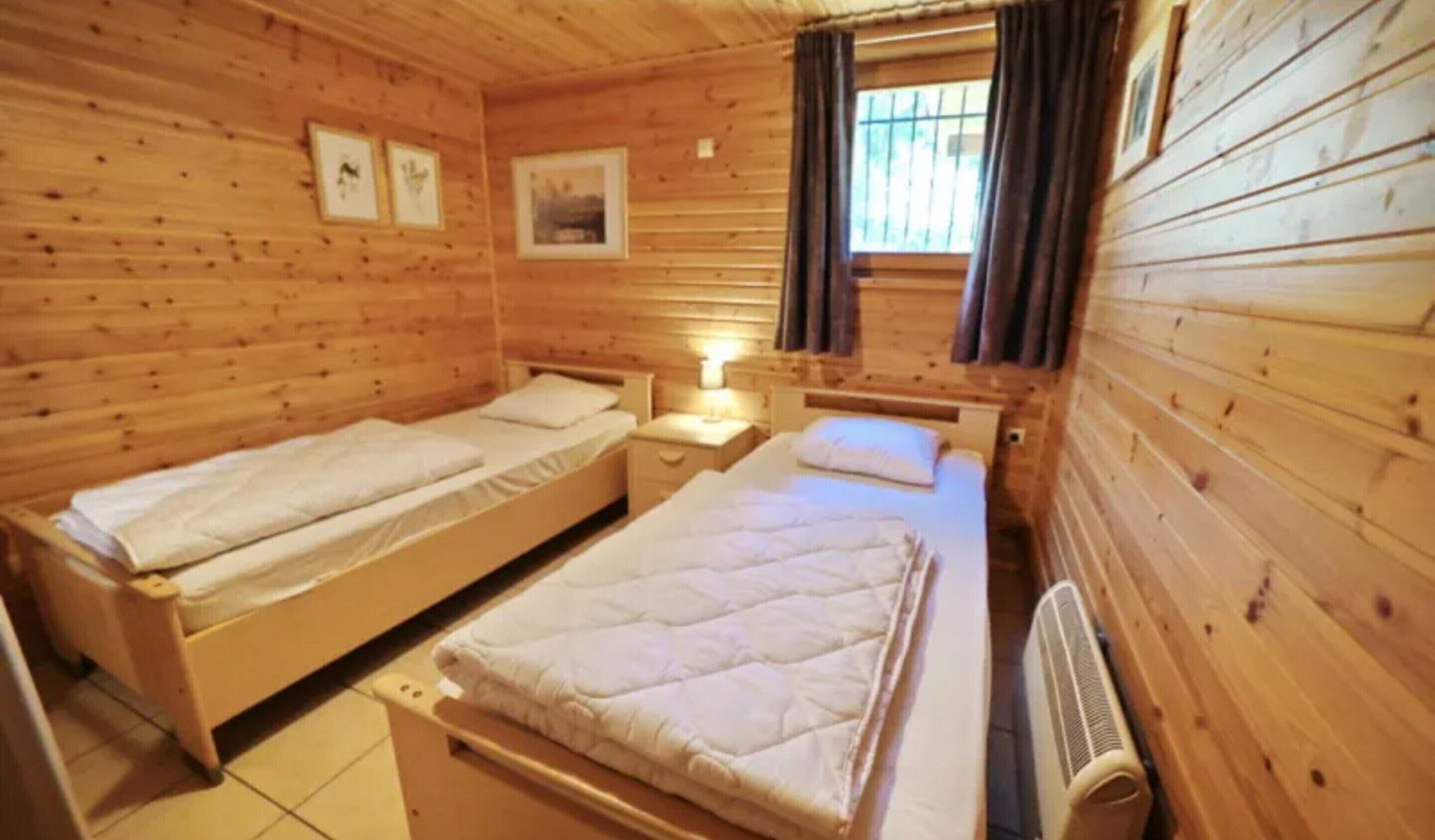 Ein Zimmer in einer Holzhütte mit zwei Einzelbetten, einem kleinen Tisch und einem Fenster mit Vorhängen.