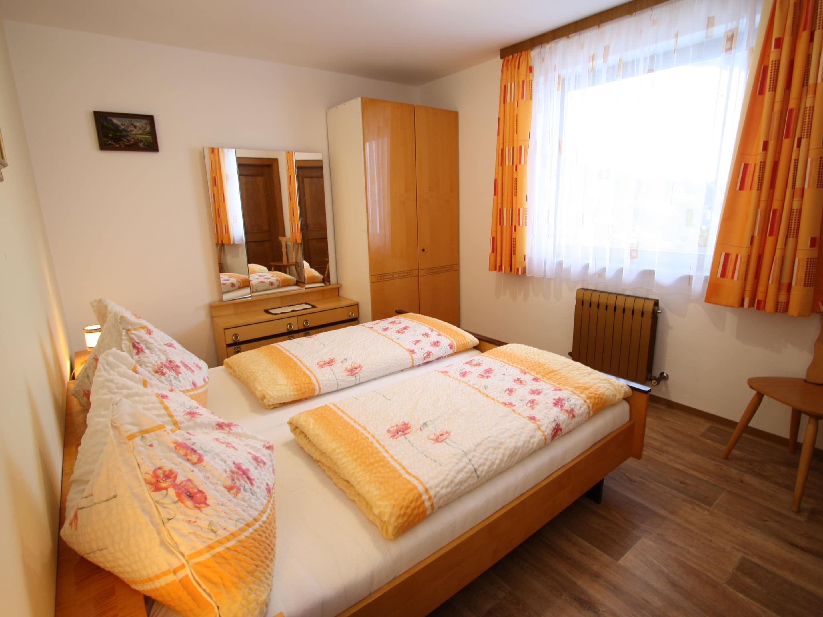 A bedroom with a double bed, wooden wardrobe, mirror, and a window with curtains.