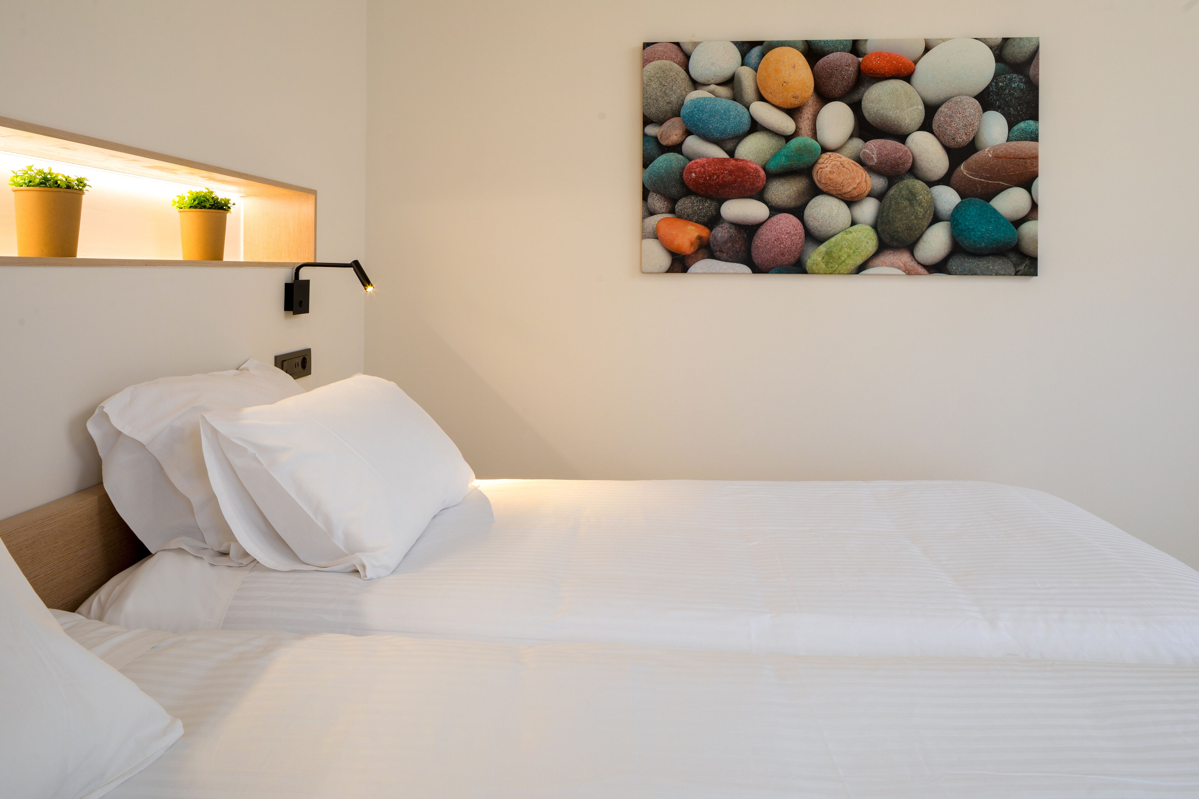 Un lit bien fait, avec du linge de lit blanc, une tête de lit et une étagère fixée au mur, sur laquelle sont posées deux plantes en pot.