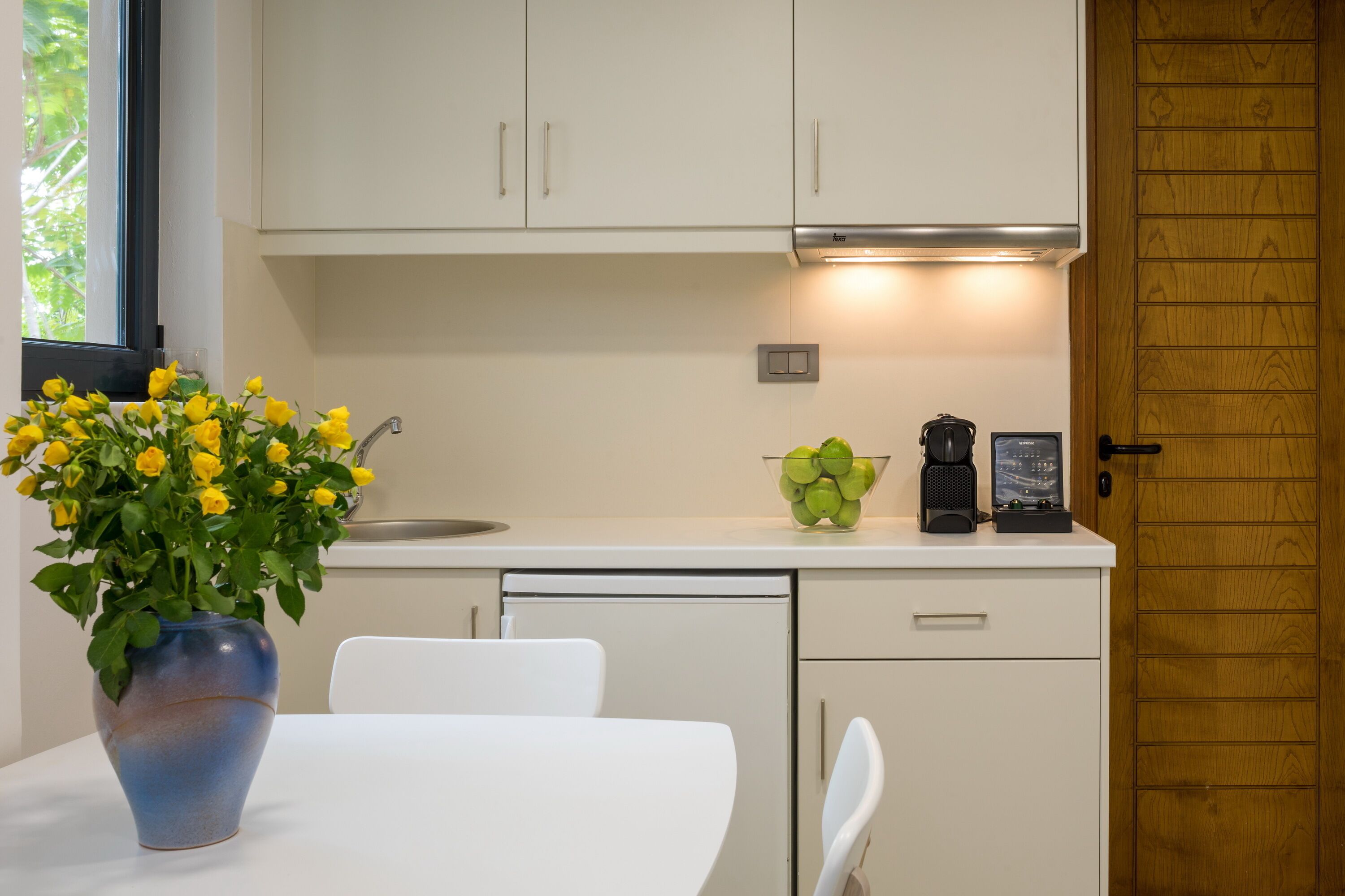 A modern kitchen with white cabinets, a white countertop, a coffee maker, and a bowl of green apples.