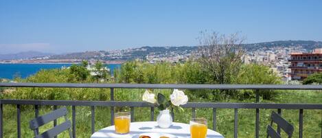 Un balcone con un tavolo apparecchiato per due persone, con un piatto di frutta e due bicchieri di succo d'arancia, con vista sulla città costiera.