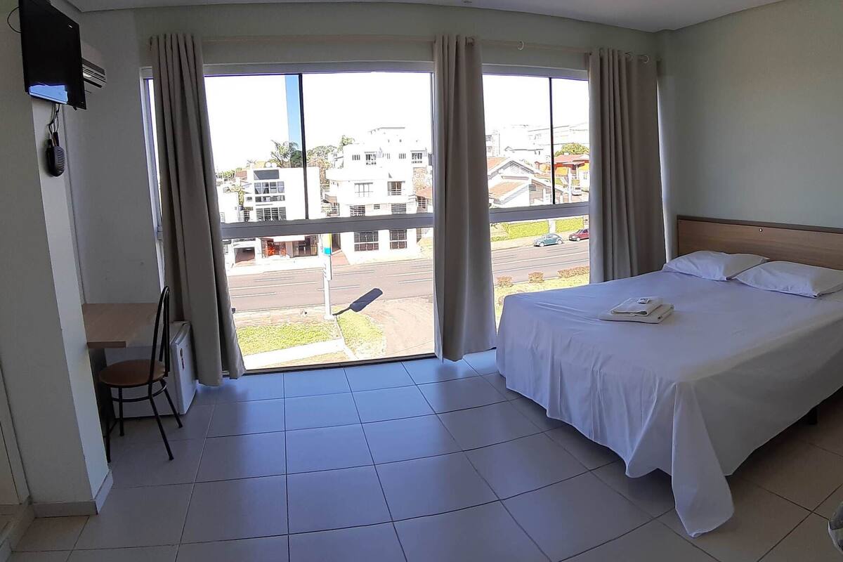 Ein Hotelzimmer mit Bett, Schreibtisch und Stuhl. Ein Fenster mit Vorhängen bietet Blick auf Gebäude und eine Straße.