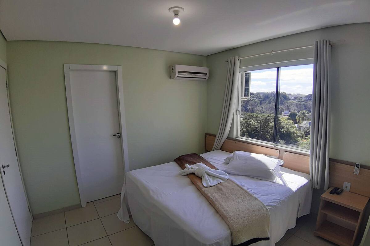 Ein Hotelzimmer mit Bett, Fenster mit Blick auf Bäume, Klimaanlage und Nachttisch mit Fernbedienung.
