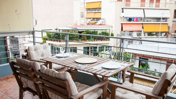 Una mesa y sillas de madera en un balcón con vistas a edificios y plantas cercanas.