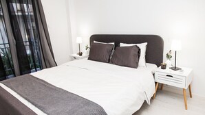 Un dormitorio moderno con una cama grande, ropa de cama blanca, dos mesitas de noche con lámparas y vistas a través de cortinas translúcidas.
