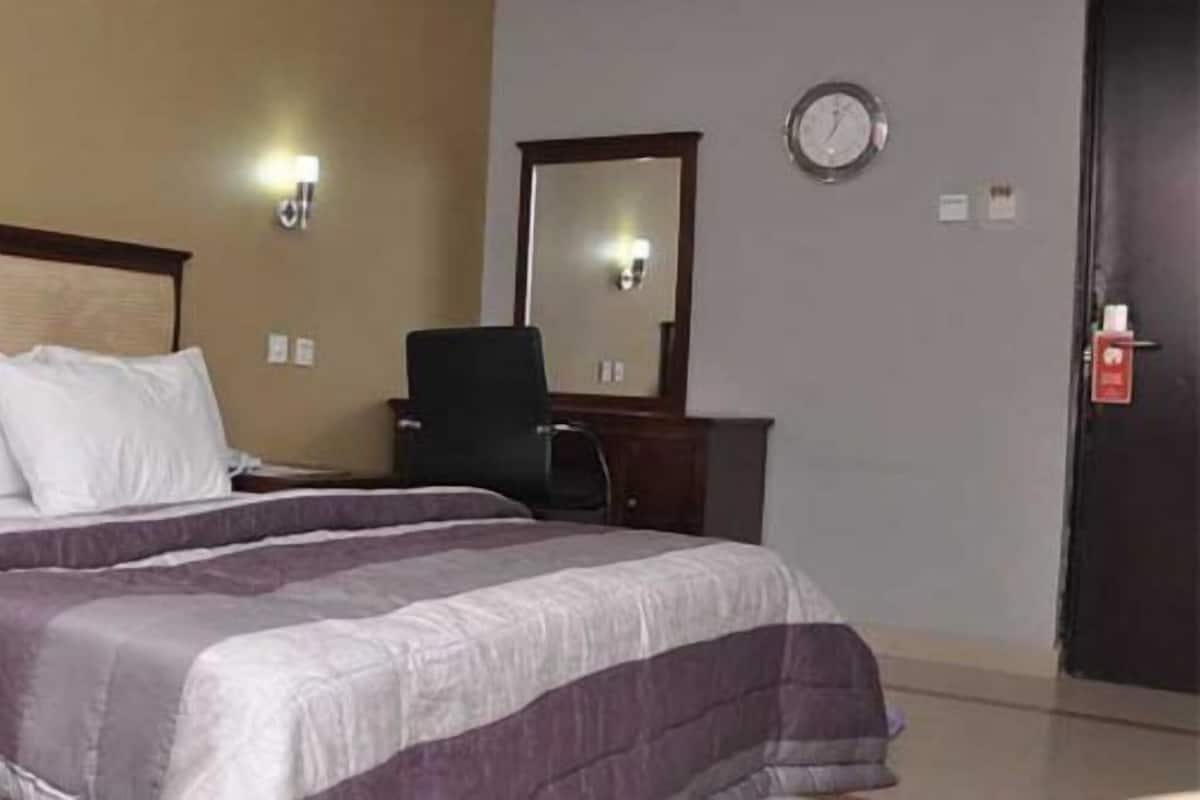 A hotel room with a bed, a chair, a desk, a mirror, and a clock on the wall.