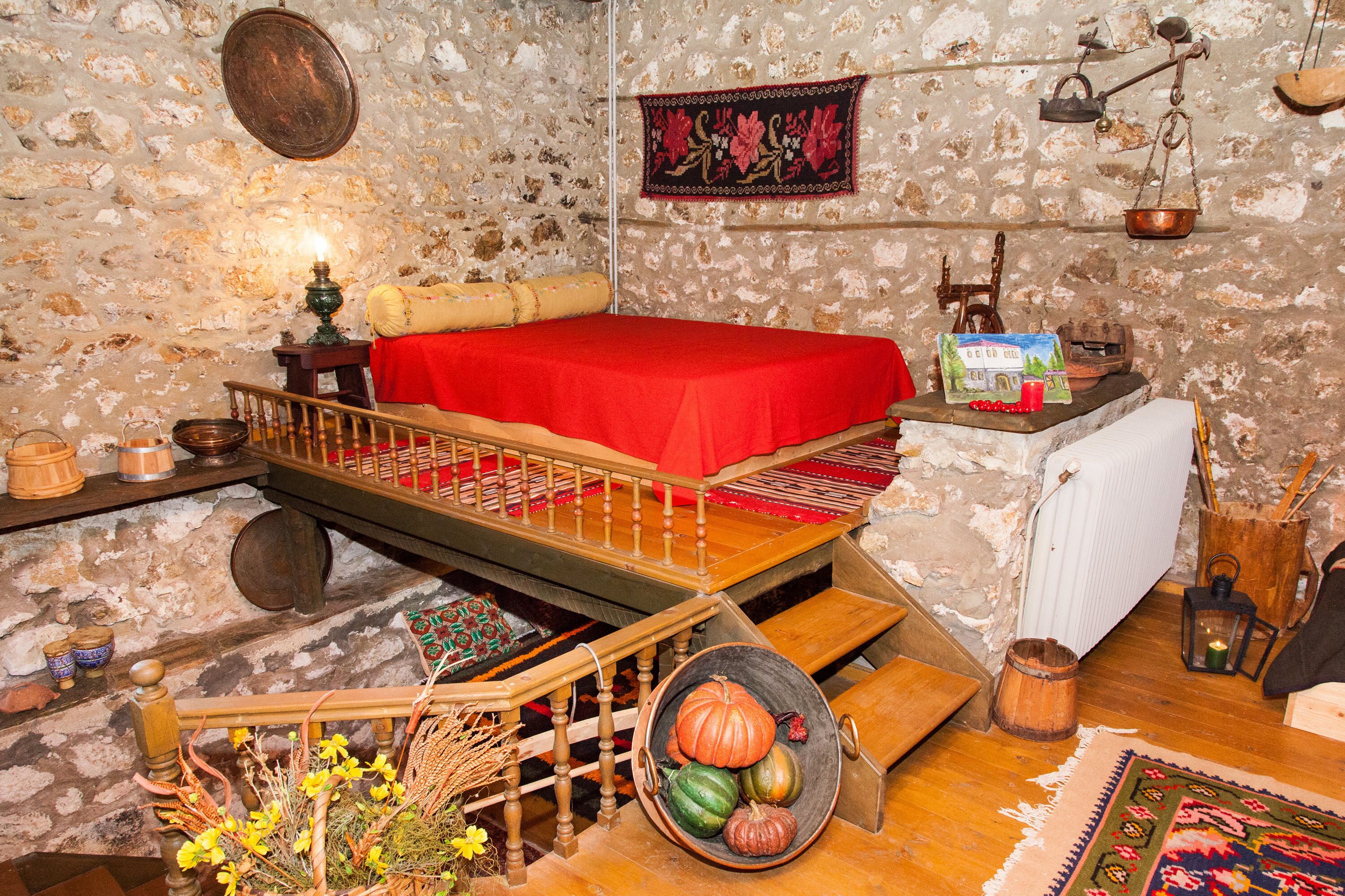A rustic room with a wooden bed, a stone wall, a red bedspread, a wooden ladder, a small table with a candle, and a hanging scale.
