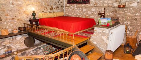 A rustic room with a wooden bed, a stone wall, a red bedspread, a wooden ladder, a small table with a candle, and a hanging scale.