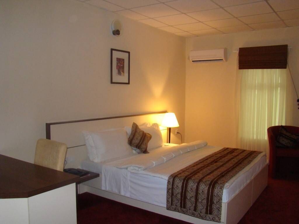 A hotel room with a bed, a desk, a chair, a lamp, and an air conditioning unit.