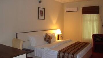 A hotel room with a bed, a desk, a chair, a lamp, and an air conditioning unit.