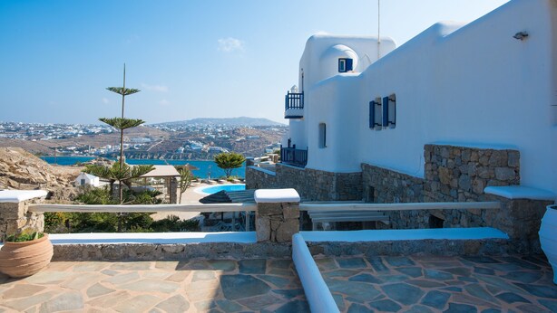 A white building with stone accents, a stone pool, and a view of a coastal town. - Villa Anemoessa Olive Green with Pool (Mykonos)