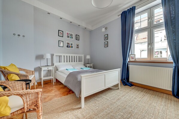 A bedroom with a white bed, wicker chairs, a bedside table, a radiator, and a window with curtains.