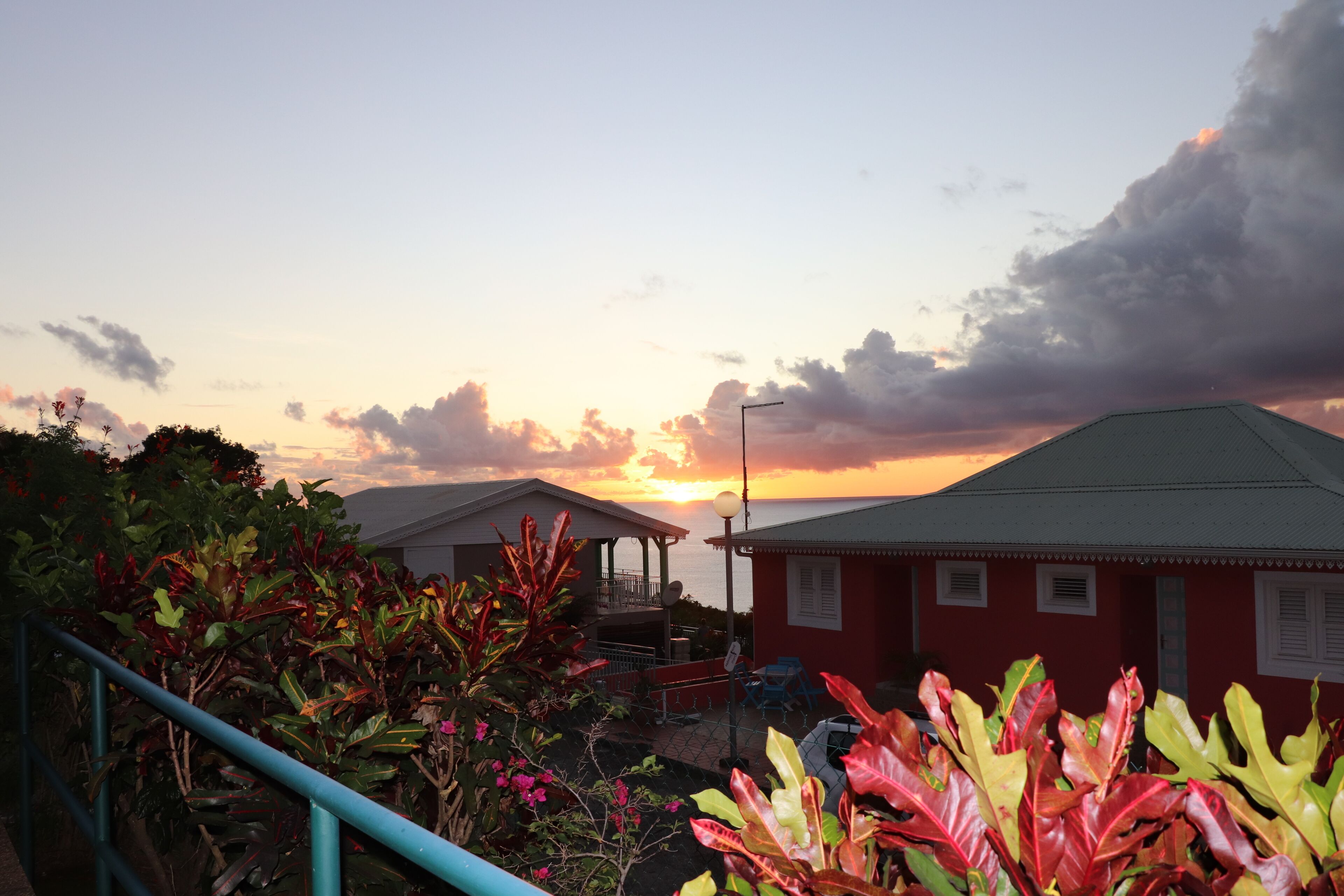 A coastal building with a red roof, surrounded by lush greenery and a sunset in the background.