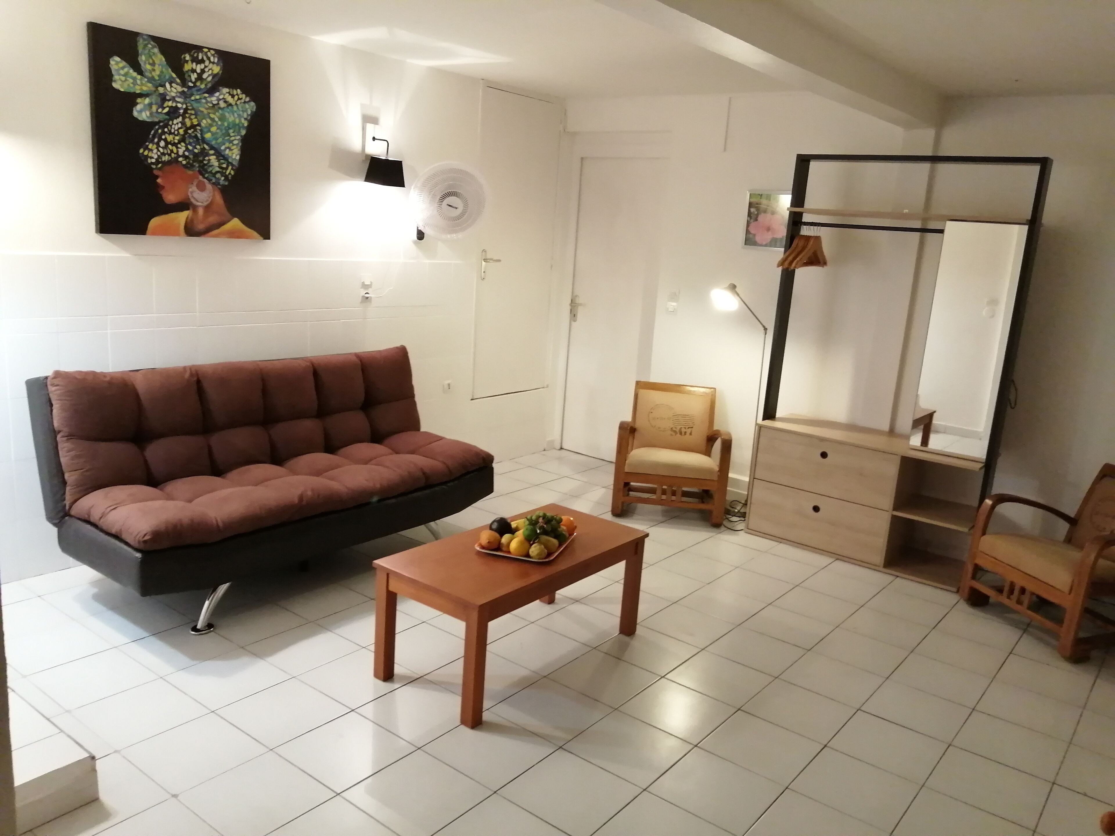 A living room with a brown sofa, a wooden coffee table with fruits, a wooden chair, a small cabinet, and a wall-mounted fan.
