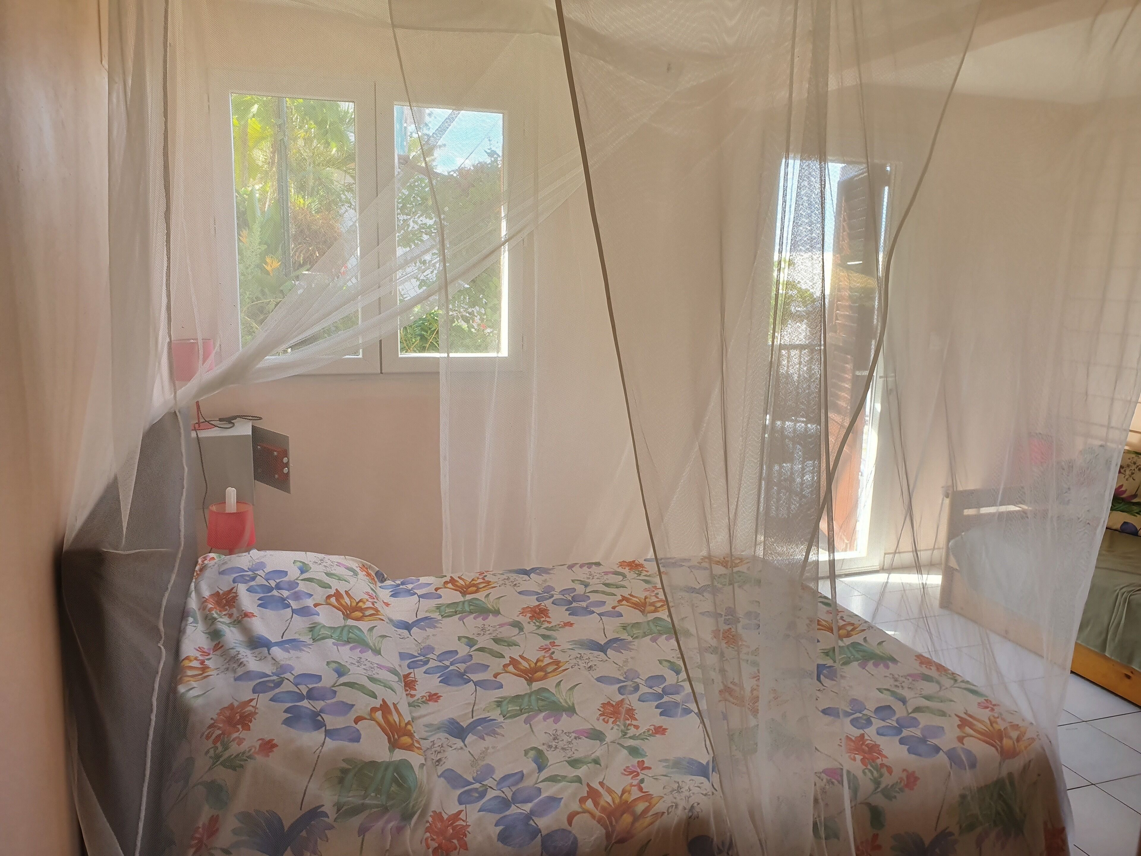 A bedroom with a bed covered by a floral bedspread, a window with a view of greenery, and a mosquito net over the bed.