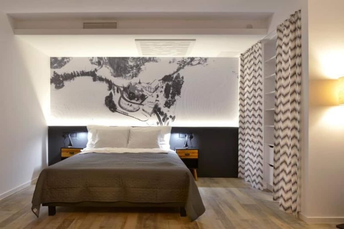 A modern bedroom with a large bed, a headboard with a wall-mounted artwork, a bedside table, a floor lamp, and patterned curtains.