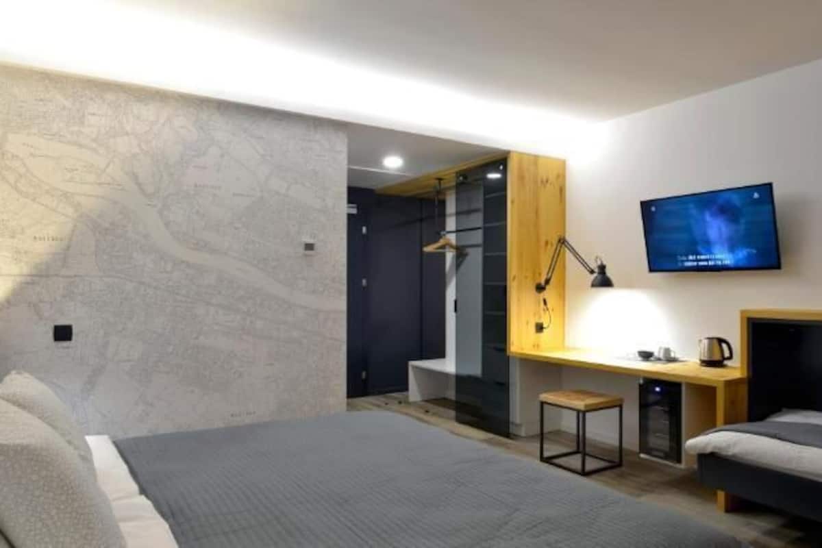 A modern hotel room with a bed, a desk with a computer, a TV, and a small stool.