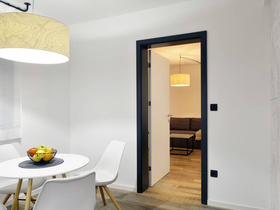 A modern dining area with a round table, white chairs, and a bowl of fruit. A pendant light hangs above the table, and a large window provides natural light.