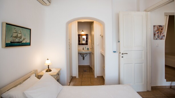 A bedroom with a bed, a nightstand with a lamp, a painting on the wall, and a door leading to a bathroom. - Villa Anemoessa Sky Blue with Pool (Mykonos)