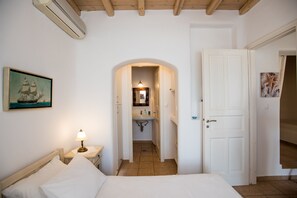 A bedroom with a bed, a nightstand with a lamp, a painting on the wall, and a door leading to a bathroom. - Villa Anemoessa Sky Blue with Pool (Mykonos)