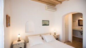 A bedroom with a bed, two bedside tables with lamps, a wall-mounted air conditioner, a painting, and an archway leading to a bathroom. - Villa Anemoessa Sky Blue with Pool (Mykonos)