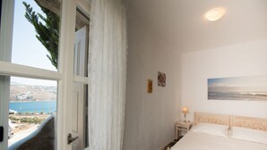 Een slaapkamer met een bed, een nachtkastje, een raam met uitzicht op de zee en een schilderij aan de muur.