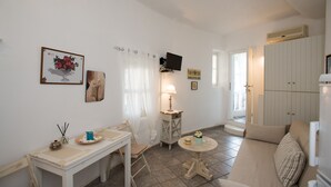 Een gezellige woonkamer met een witte bank, een kleine ronde tafel, een witte tafel met een stoel, en een televisie die aan de muur is bevestigd.