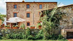 Un bâtiment en pierre doté de nombreuses fenêtres, un patio avec un parasol et des plantes en pot.