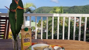 Una mesa con una bandeja de comida, un cartón de jugo y dos tazas, en una terraza de madera con vista panorámica y un adorno colgante.