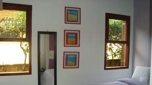 A bedroom with a bed, three framed pictures, a mirror, and two windows with views of trees. - Beautiful comfortable house (São Sebastião)