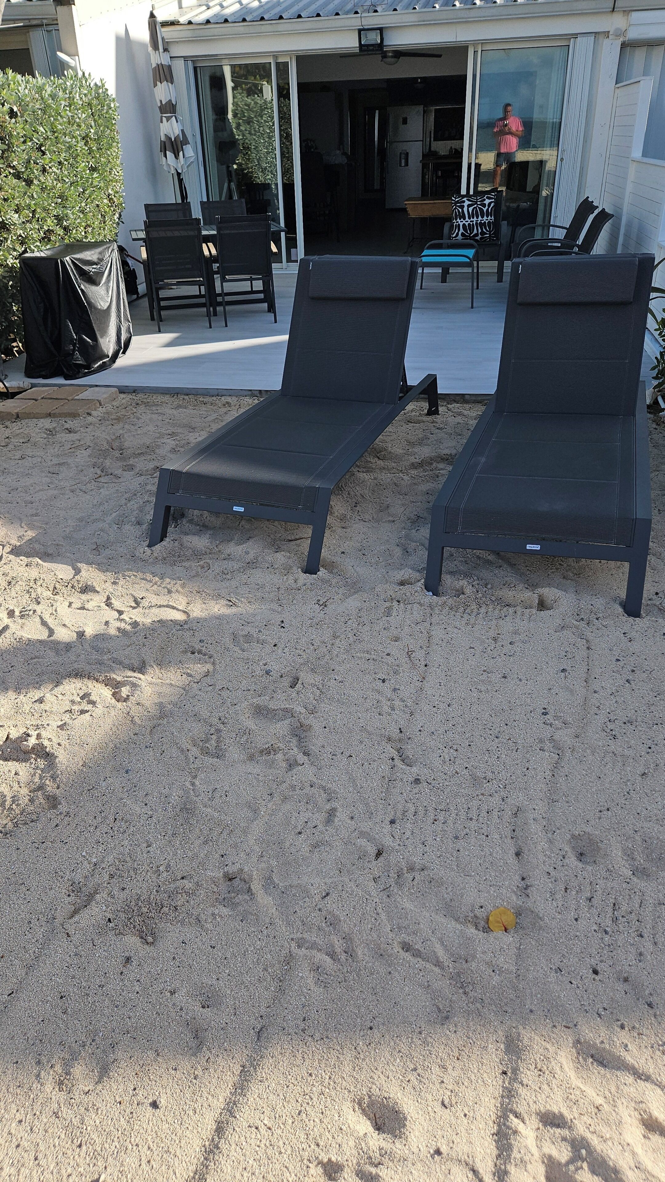 Two dark-colored lounge chairs on a sandy surface, with a covered table and chairs in the background.