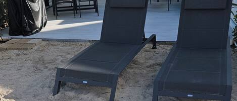 Two dark-colored lounge chairs on a sandy surface, with a covered table and chairs in the background.