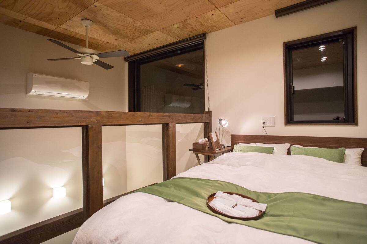 A hotel room with a wooden ceiling, a ceiling fan, a large window, a bed with green bedding, and a bedside table with a lamp.
