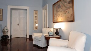 A room with a white door, two white armchairs, a round table with a lamp, and a large framed painting on the wall.