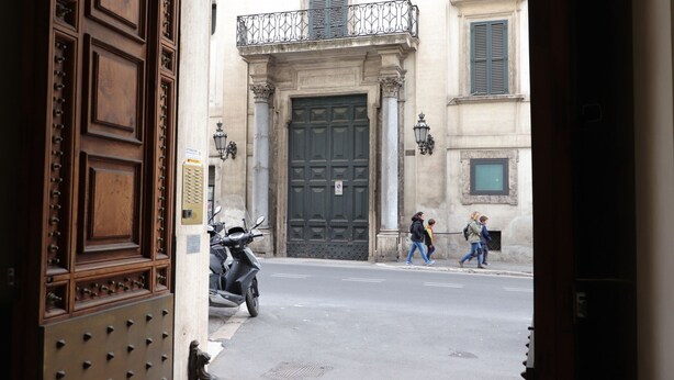 A street view with a large wooden door, a motorcycle parked on the side, and pedestrians walking on the sidewalk.