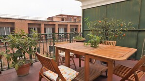 A wooden table and chairs on a balcony with potted plants and a view of buildings.