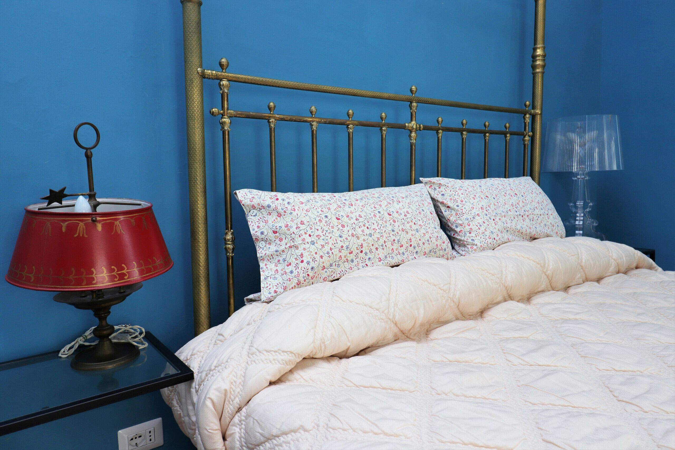 Uma cama com colcha branca, duas almofadas florais, um abajur vermelho e uma cabeceira de metal, tudo diante de uma parede azul.