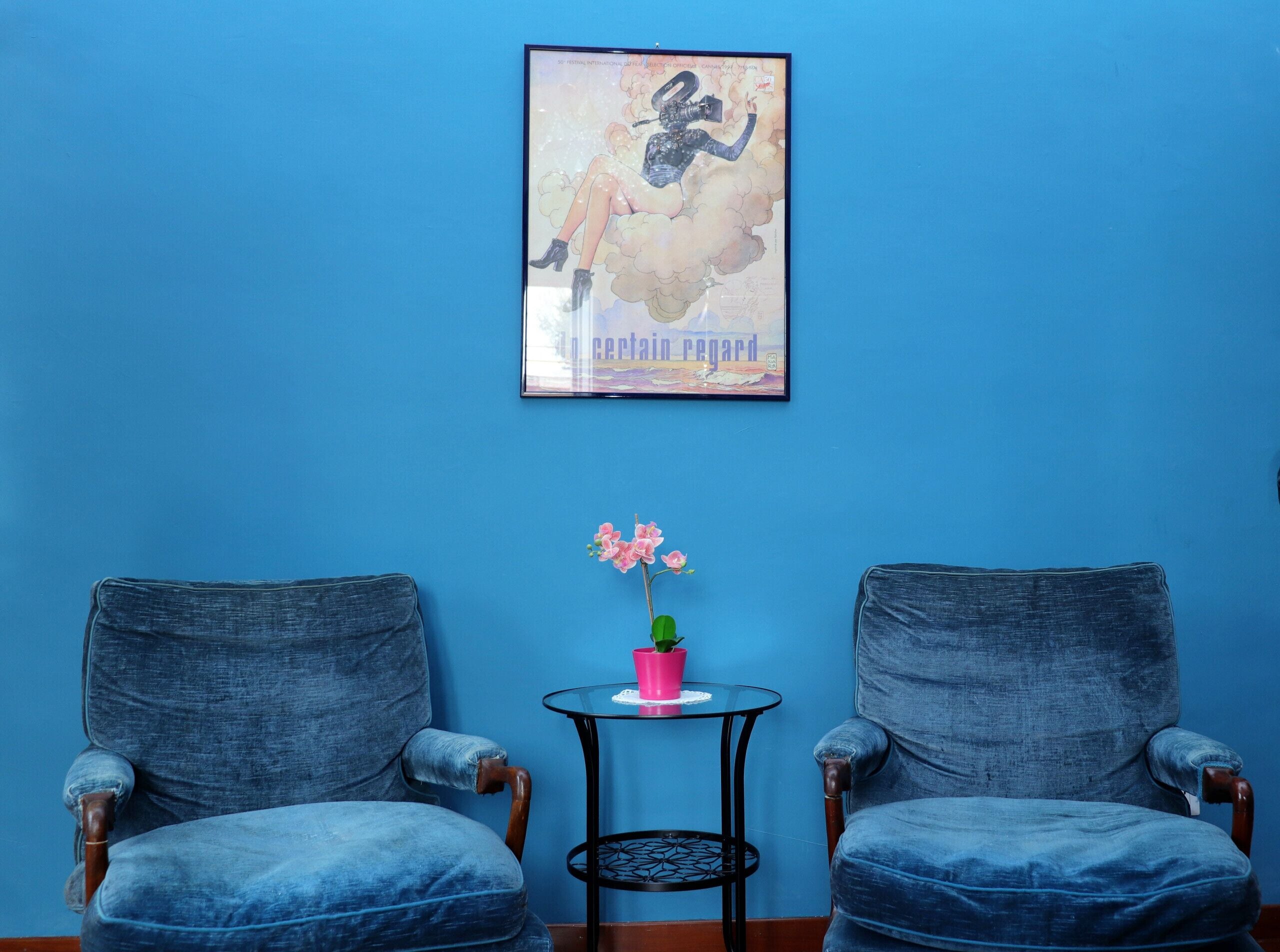A room with two blue armchairs, a small round table with a pink flower vase, and a framed poster on the wall.