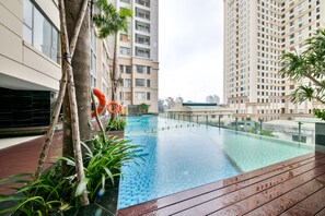 A rooftop swimming pool with a wooden deck, surrounded by tall residential buildings and greenery.