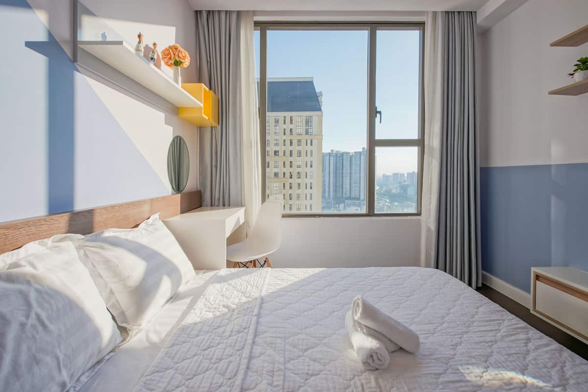 Une chambre d’hôtel moderne, dotée d’un grand lit, d’un bureau, d’une chaise et offrant une vue sur la ville grâce à une grande fenêtre.