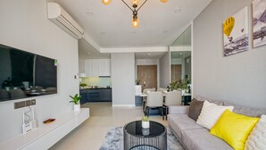 Ein modernes Wohnzimmer mit einem Flachbildfernseher, einer Couch mit gelb-weißen Kissen, einem runden Couchtisch und einem Essbereich im Hintergrund.