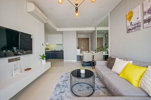 Uma sala de estar moderna com uma televisão de tela plana, um sofá com almofadas amarelas e brancas, uma mesa de centro redonda e uma área de jantar ao fundo.